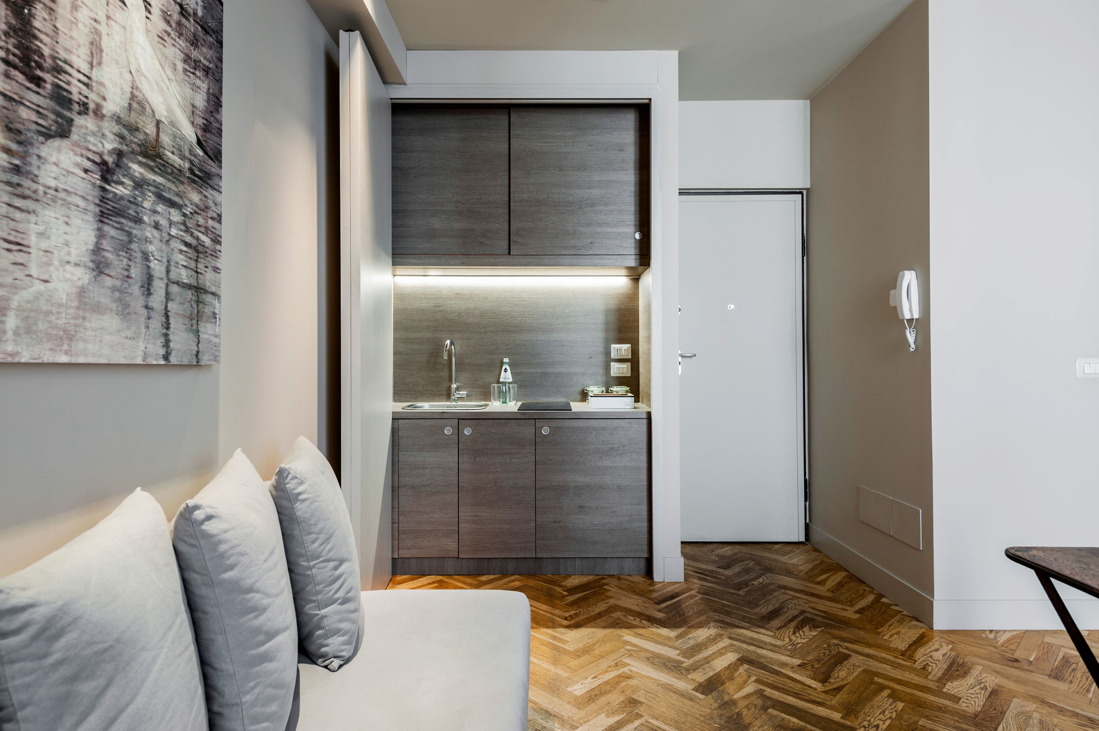 Un espacio habitable compacto con kitchenette, un sofá con cojines y un suelo de madera con patrón de espiga.