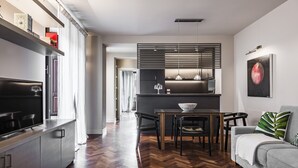 A modern living room with a sofa, a dining table with chairs, a kitchen area with a counter and appliances, and a hallway leading to another room. - Ferrini Home - Via Monte Sant'Agata (Catania)