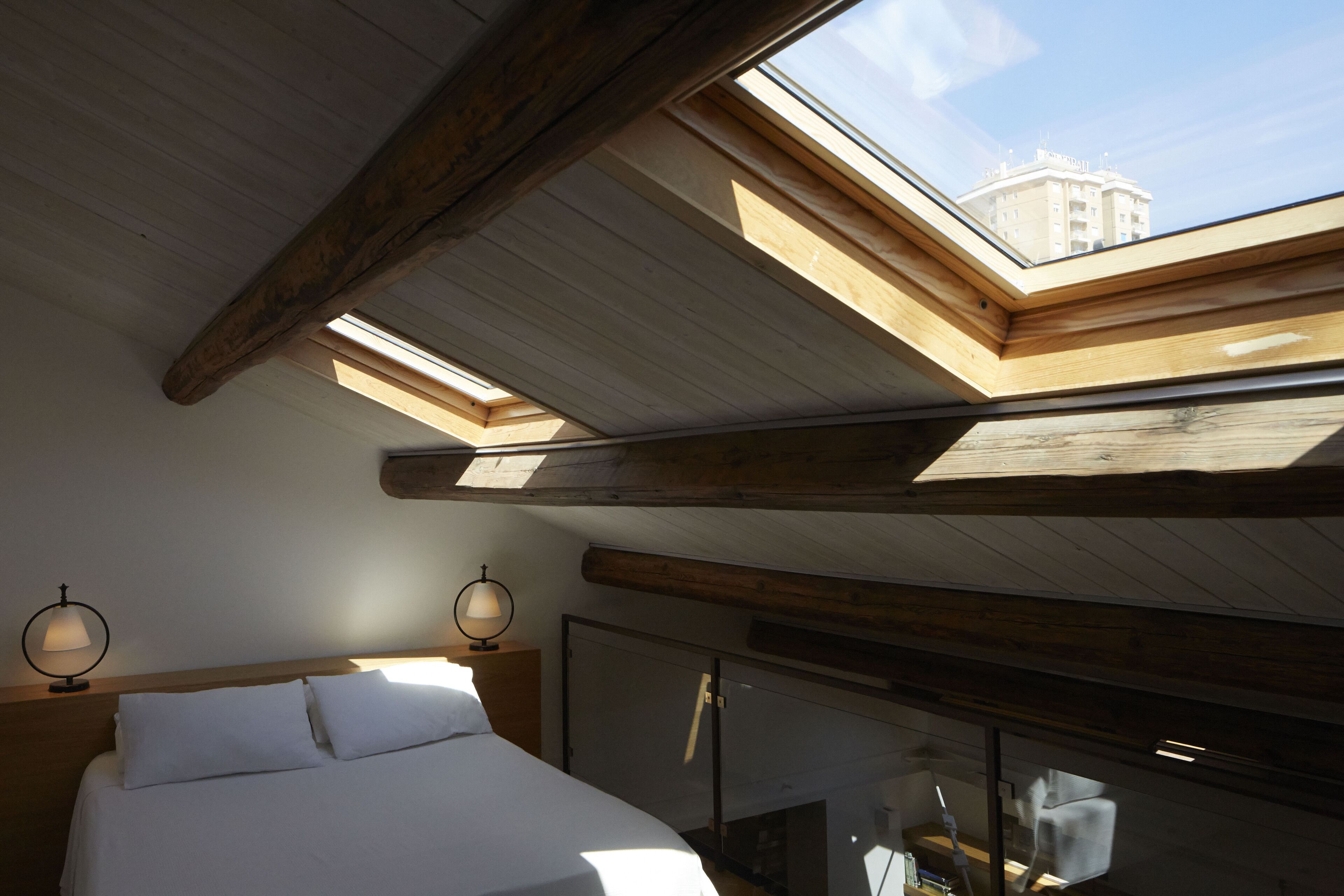 A bedroom with a skylight, wooden beams, a bed, and two wall-mounted lamps.