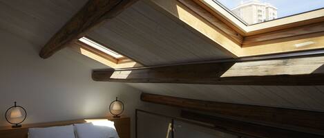 A bedroom with a skylight, wooden beams, a bed, and two wall-mounted lamps.