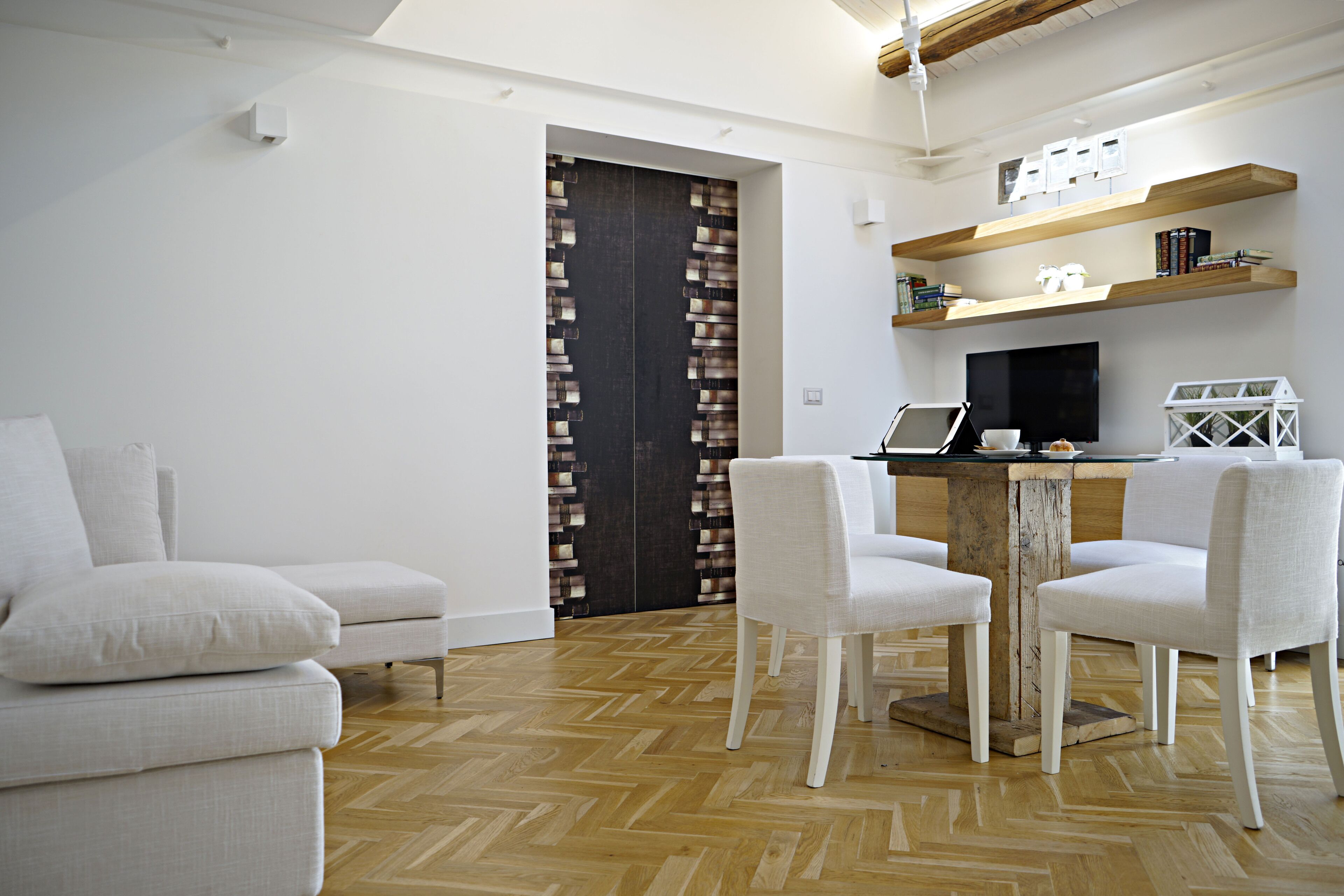 A modern living room with a herringbone wood floor, a sofa, a dining table with chairs, and a dark wooden door with a patterned design.