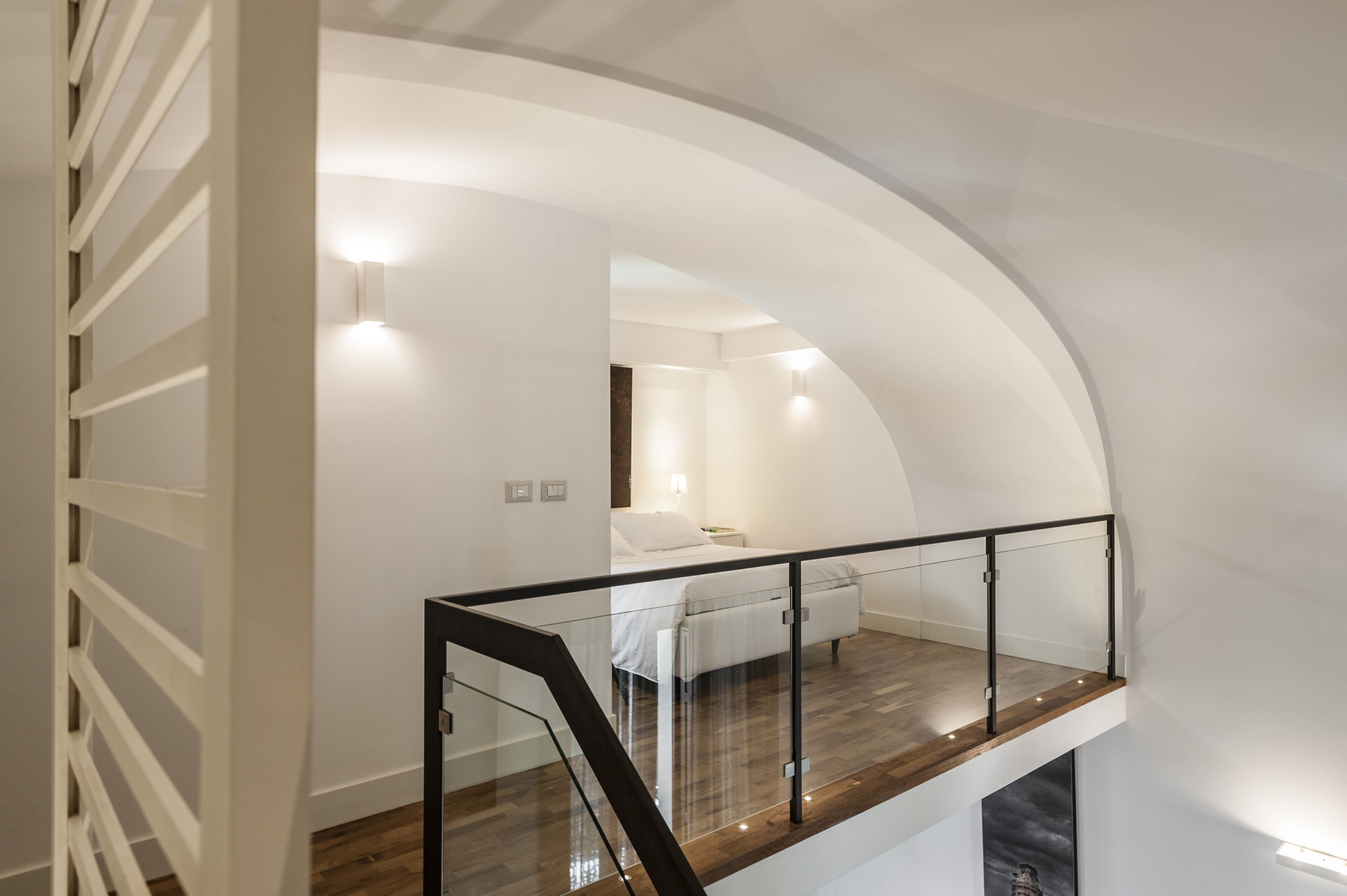 A modern interior with a curved staircase, a bedroom with a bed and bedside table, and a glass railing on the upper floor.
