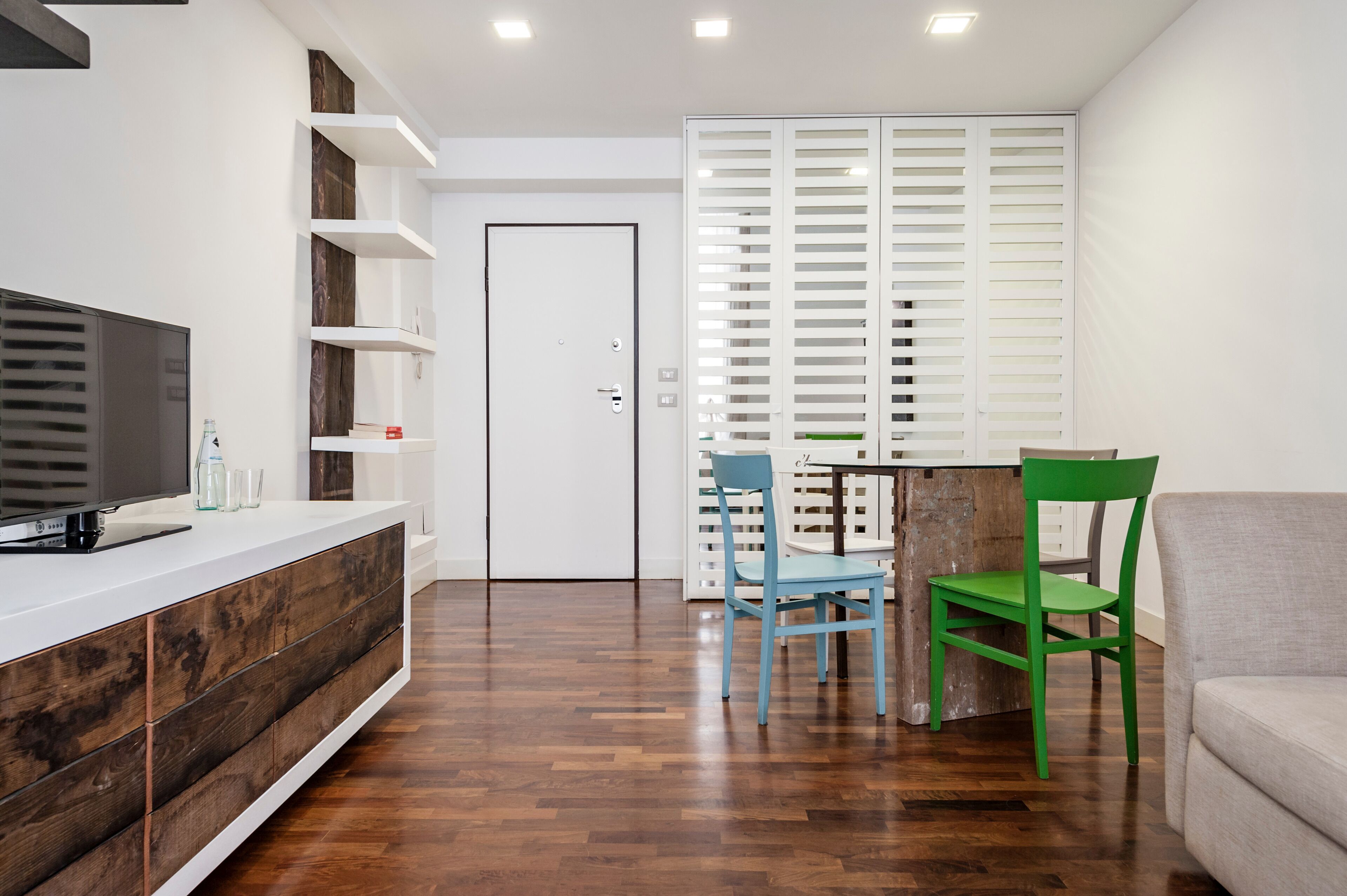 Una sala moderna con televisor de pantalla plana, mueble de almacenamiento de madera y una zona de comedor con dos sillas.