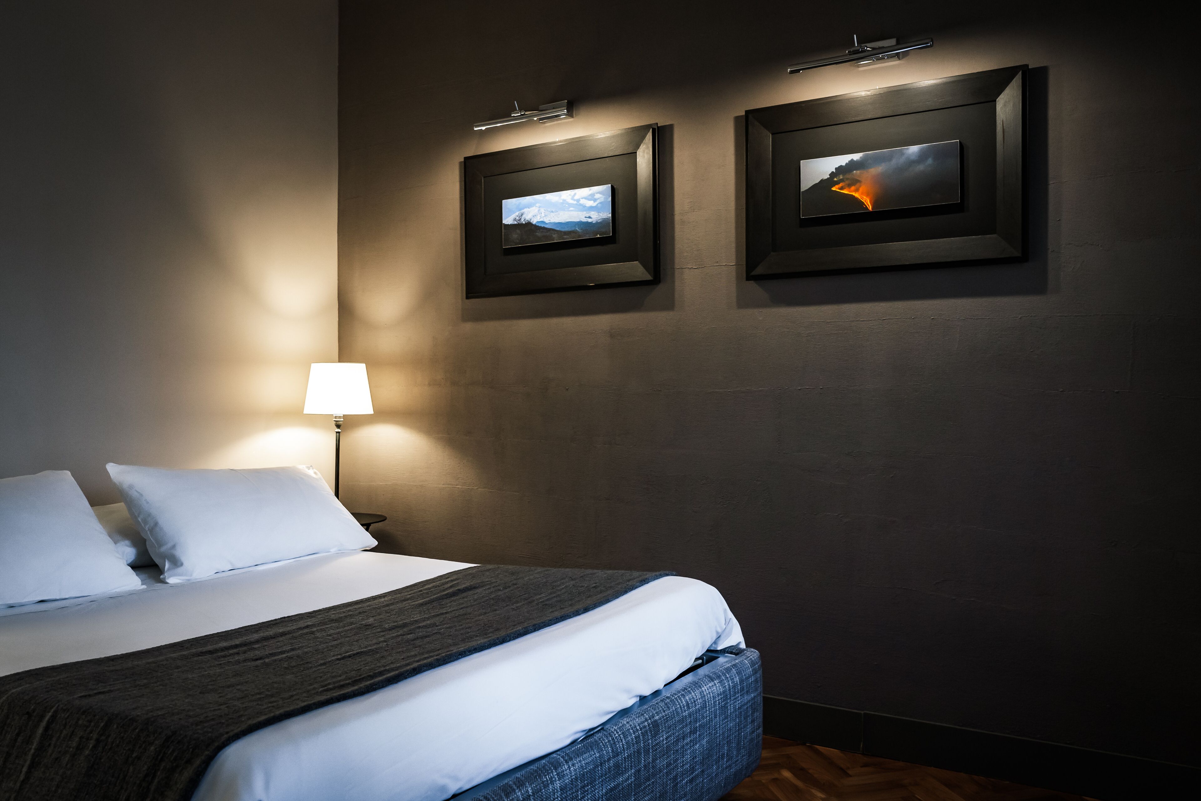 Hotellihuone, jossa on siististi pedattu sänky, lamppu ja kaksi kehystettyä kuvaa seinällä.