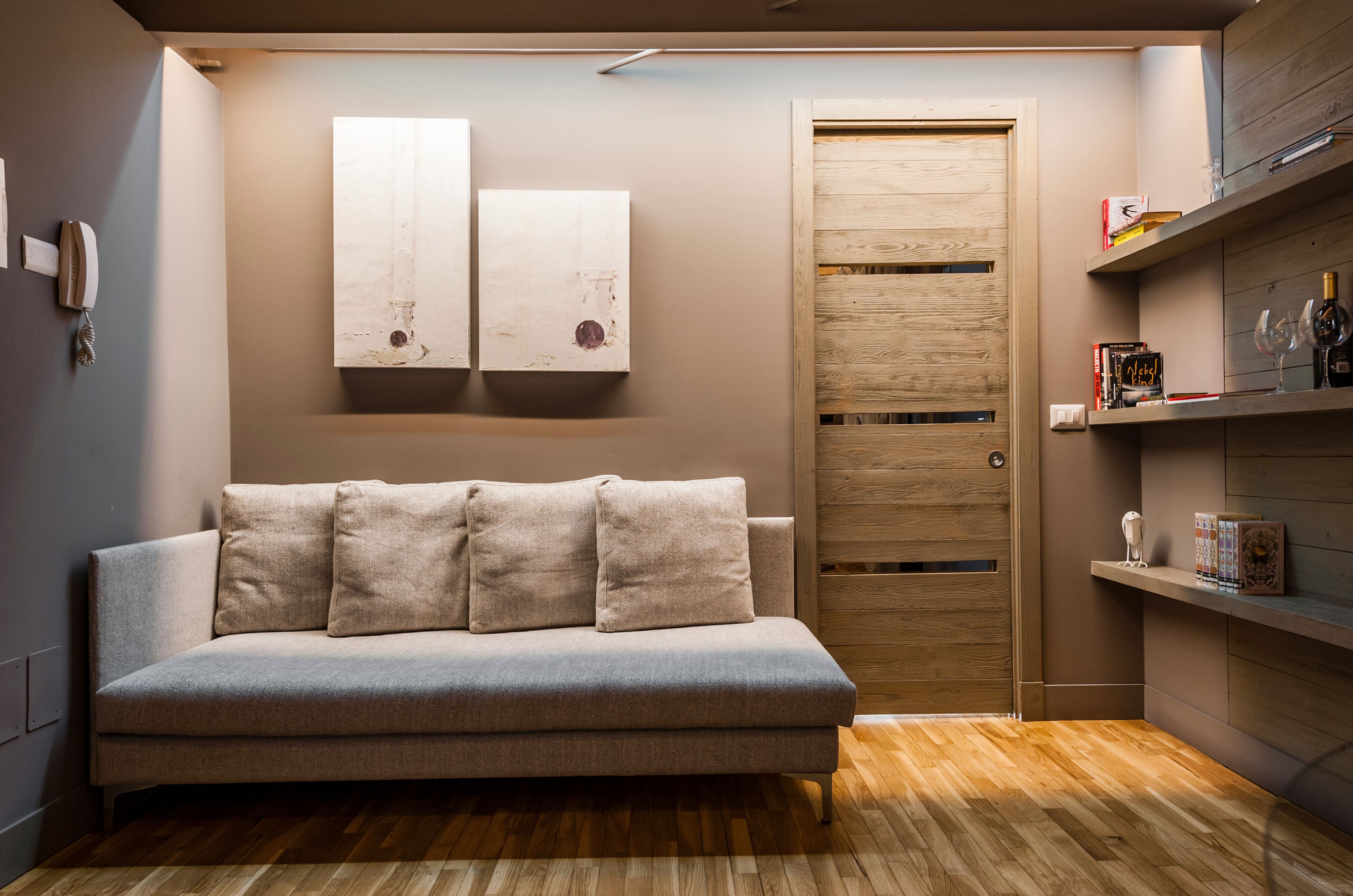 A modern living room with a grey sofa, wooden door, and shelves with books and decorative items.