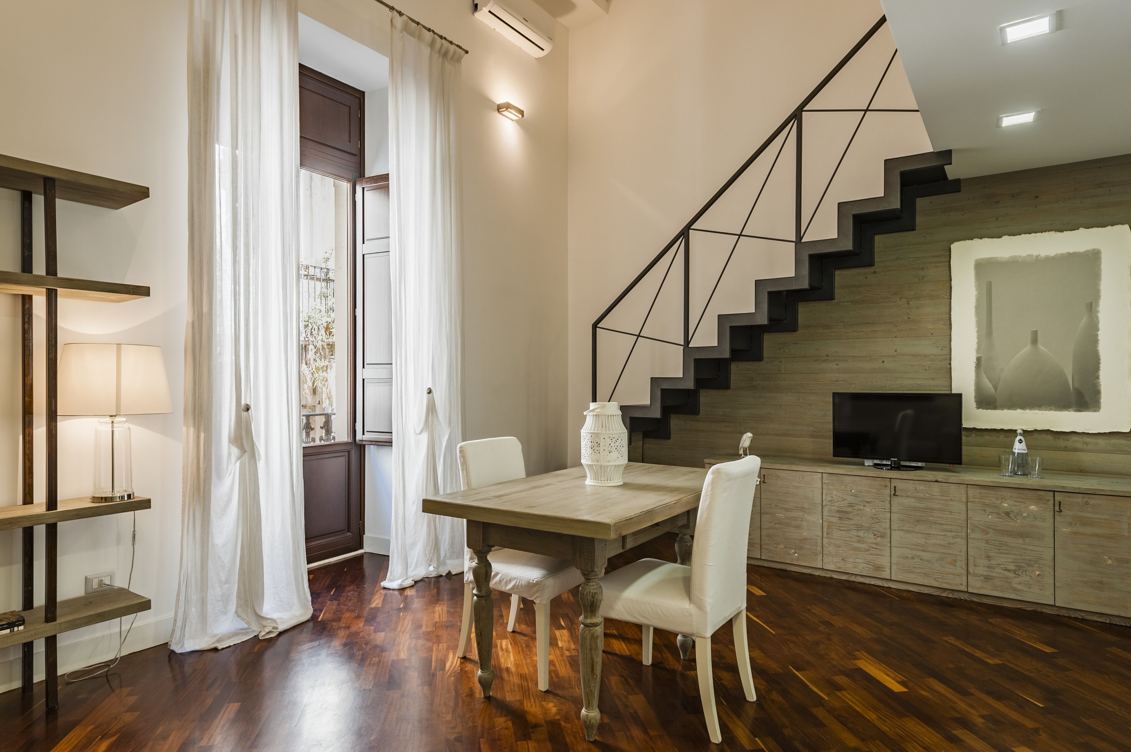 A modern living room with a wooden dining table, white chairs, a TV, and a staircase with a geometric railing.