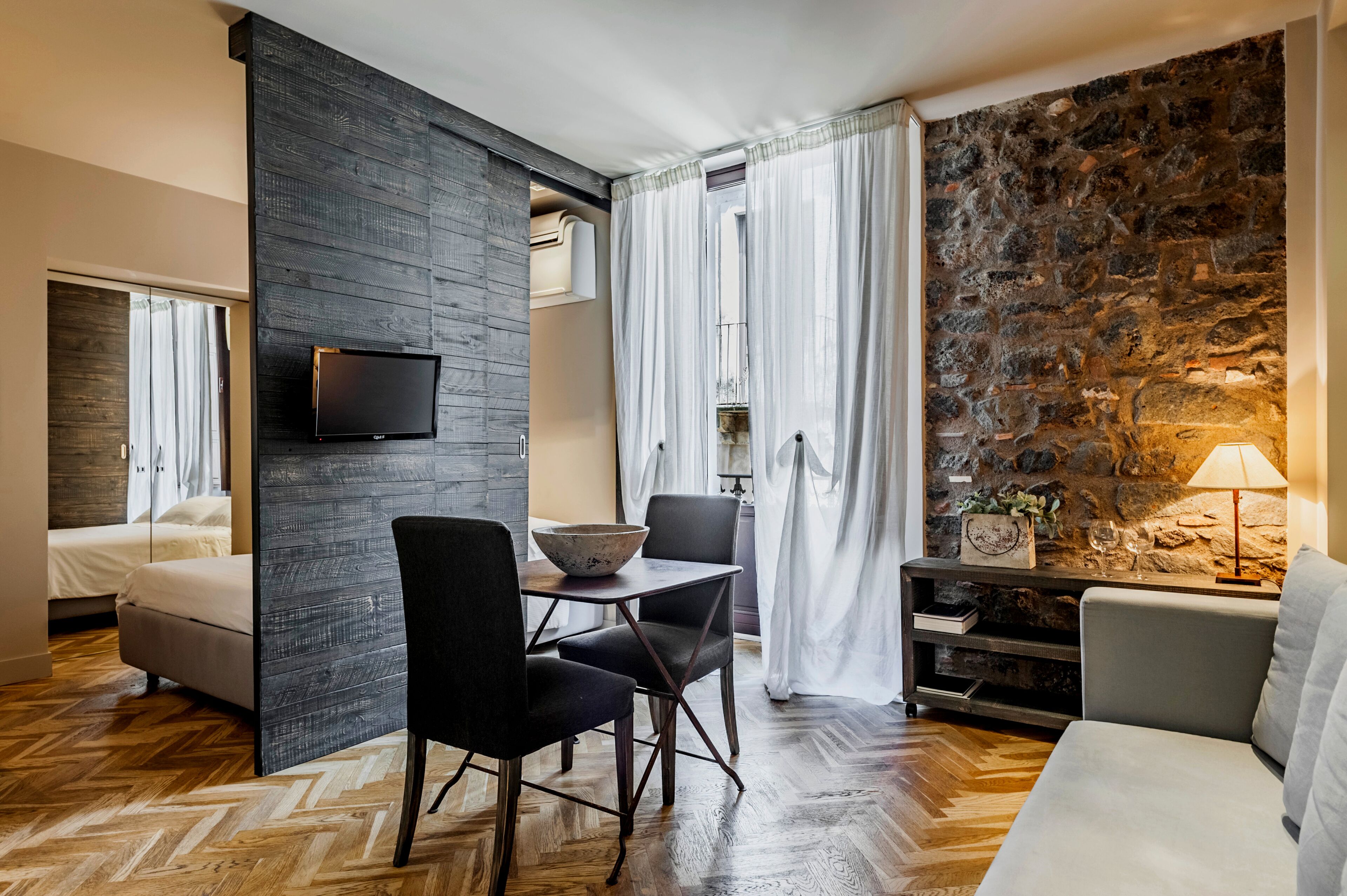 Ett modernt rum med en vägg av sten, en platt-TV, ett matbord med stolar, en soffa och en lampa.