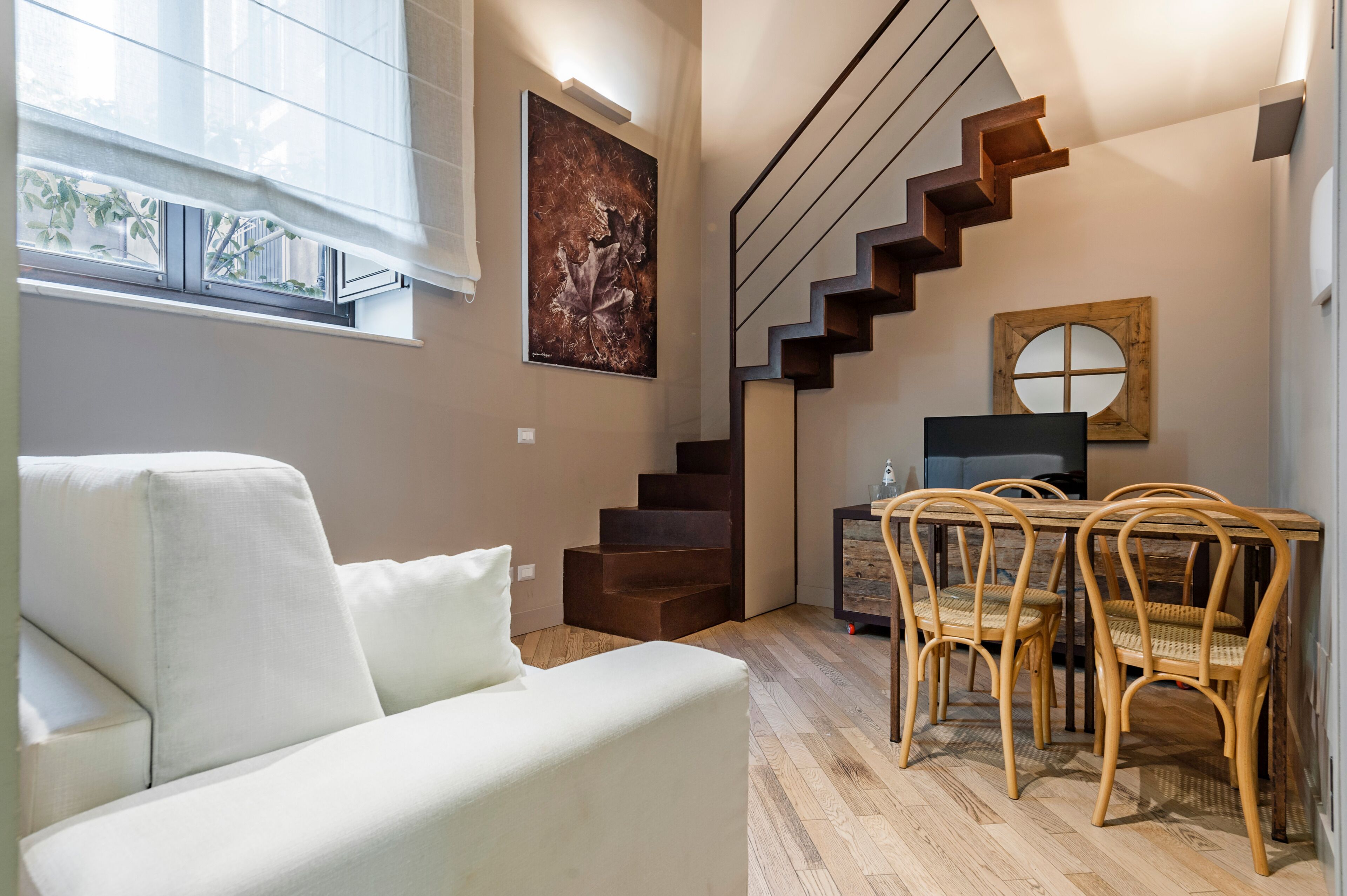 Una sala moderna con sofá, escalera de madera, mesa de comedor con sillas y un cuadro en la pared.