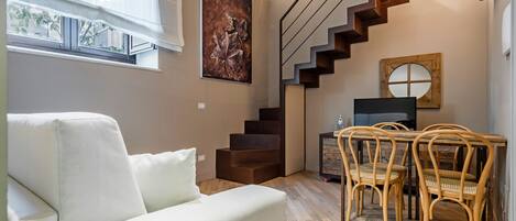 A modern living room with a sofa, wooden staircase, dining table with chairs, and a painting on the wall.