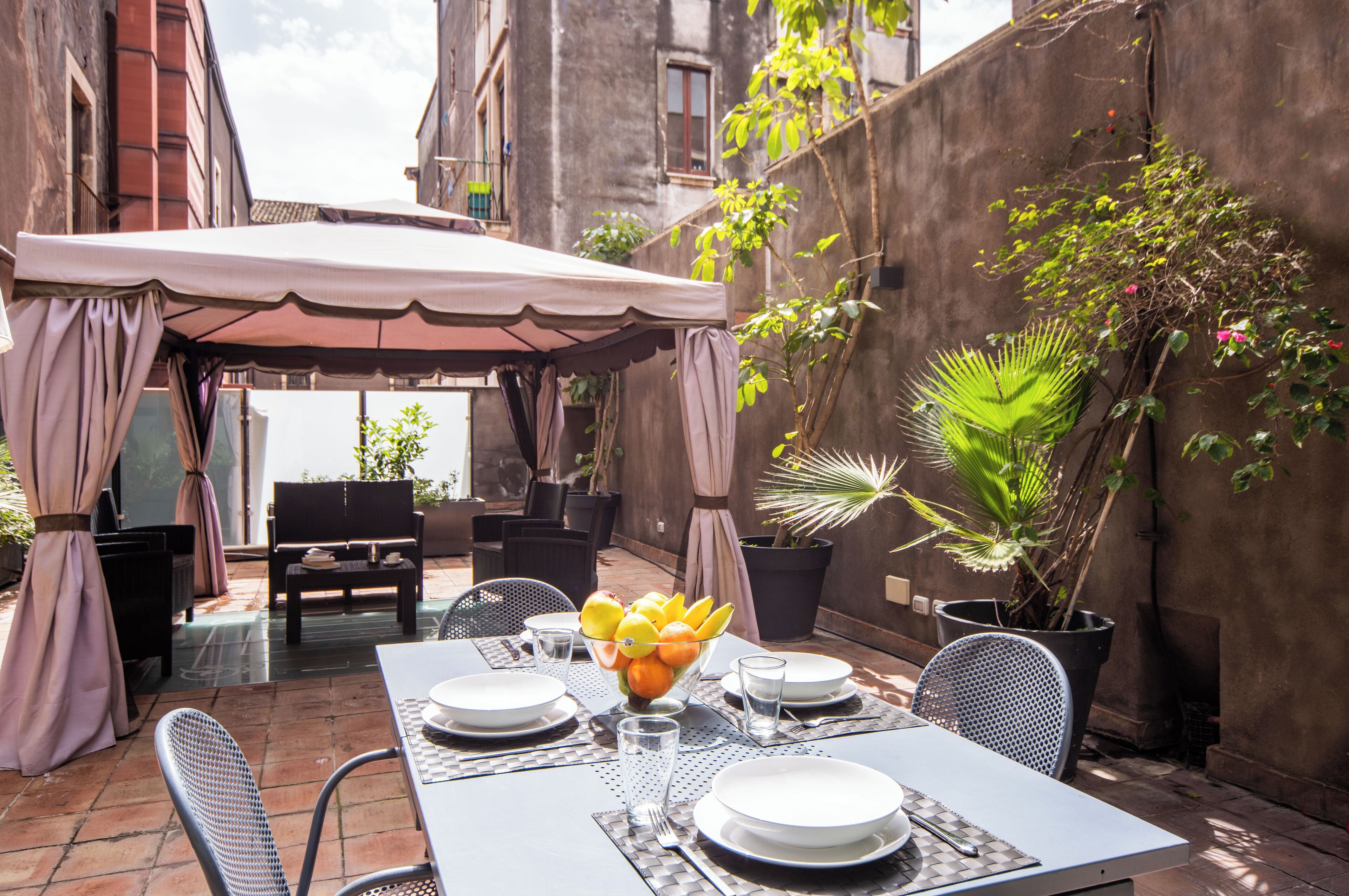 Un área de comedor al aire libre con una mesa preparada para cuatro personas, rodeada de sillas, bajo un toldo.