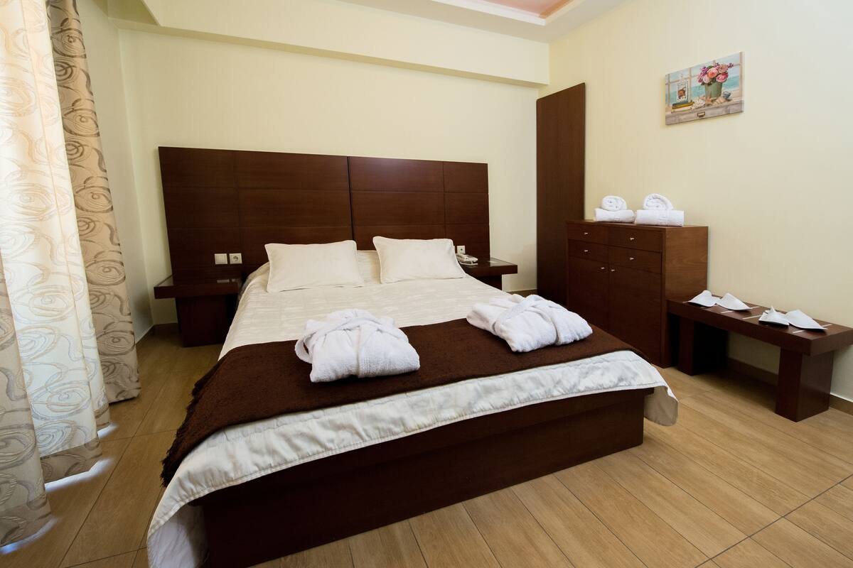 A neatly made bed with white linens and towels, a wooden headboard, a nightstand with a phone, and a wooden cabinet with towels.