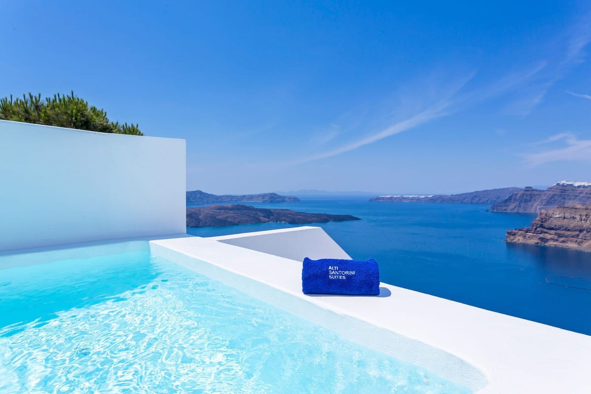Una piscina azul cristalina con una toalla que dice "ALTI SANTORINI SUITES" y vistas al mar y a los acantilados.