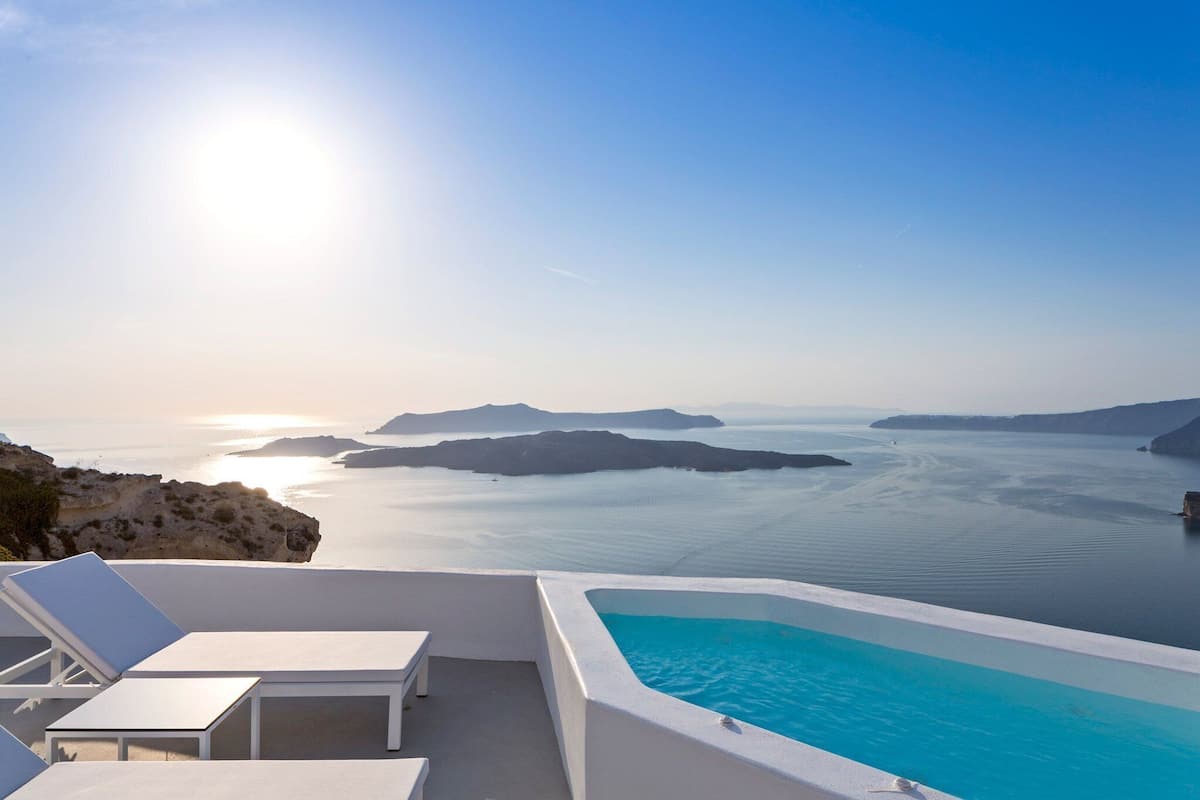 Una piscina infinita iluminada por el sol, con sillas de descanso blancas y vistas al mar y a las islas.