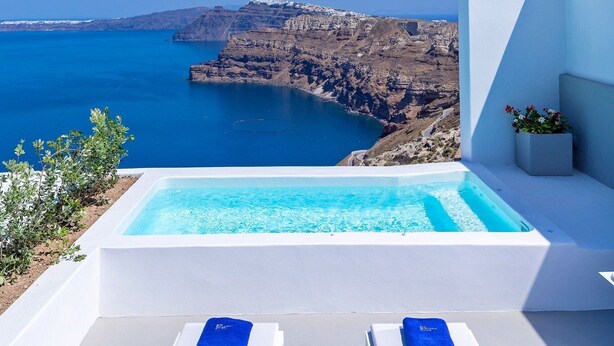 A modern outdoor pool area with two lounge chairs and a small table, overlooking a cliffside and the sea. - Alti Santorini Suites (Santorini)