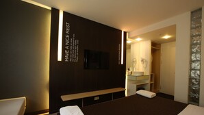 A modern hotel room with a large bed, a bedside table, a flat-screen TV, a built-in sink, and a mirror.