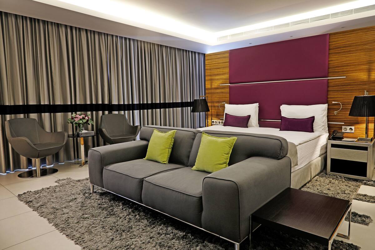 A modern hotel room with a grey sofa, a bed with white and purple bedding, and a wooden accent wall.