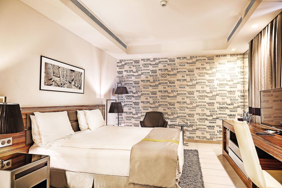 A hotel room with a bed, bedside table, lamp, and a wall with city names.