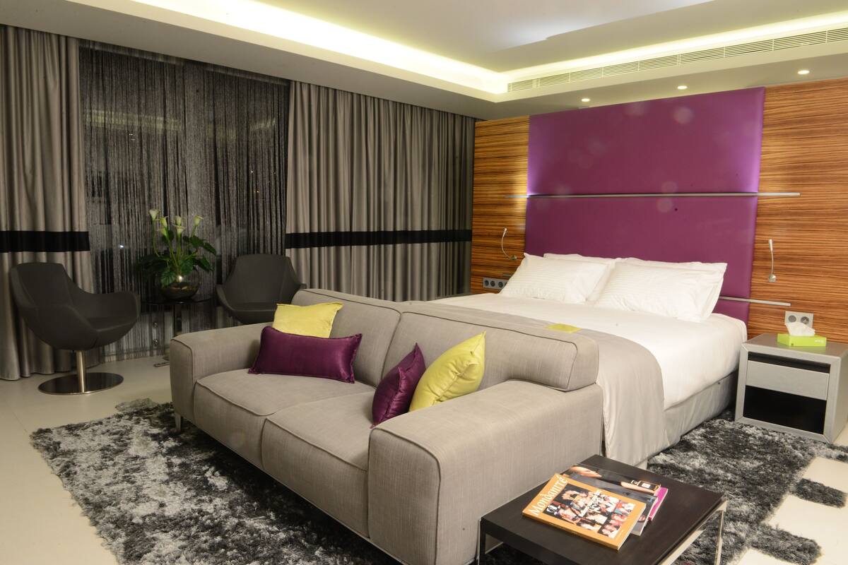 A modern hotel room with a large bed, a grey sofa with colorful cushions, a small side table, and a black coffee table.