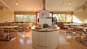 A modern dining area with round tables and chairs, a central serving station, and large windows with a view of trees. - Contact Hotel du Parc (Orvault)