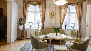 A room with a round table, six upholstered chairs, a chandelier, two windows with curtains, and a potted plant on a side table.