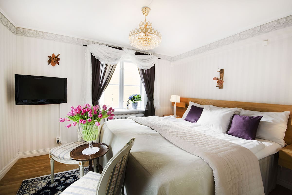 Una habitación de hotel con una cama grande, un televisor de pantalla plana montado en la pared, una araña y una ventana con cortinas.