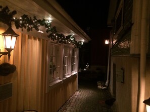 A narrow cobblestone alleyway with wooden buildings on either side, adorned with string lights and a lantern on the left.