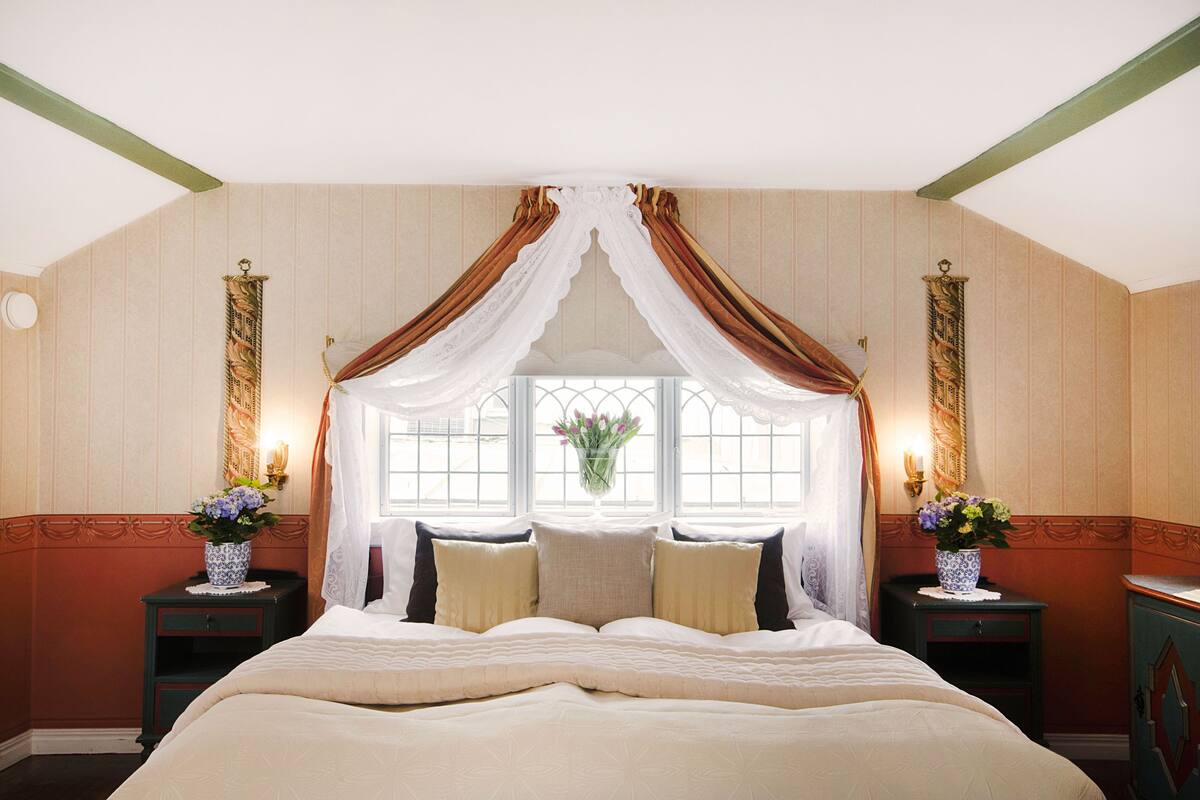 Un dormitorio con una cama grande, una ventana con cortinas, dos mesitas de noche con lámparas y un estante en la pared con flores.