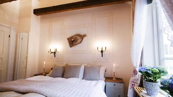 A neatly made bed with a white duvet, two bedside tables with candles, a wall-mounted clock, and a window with a curtain.