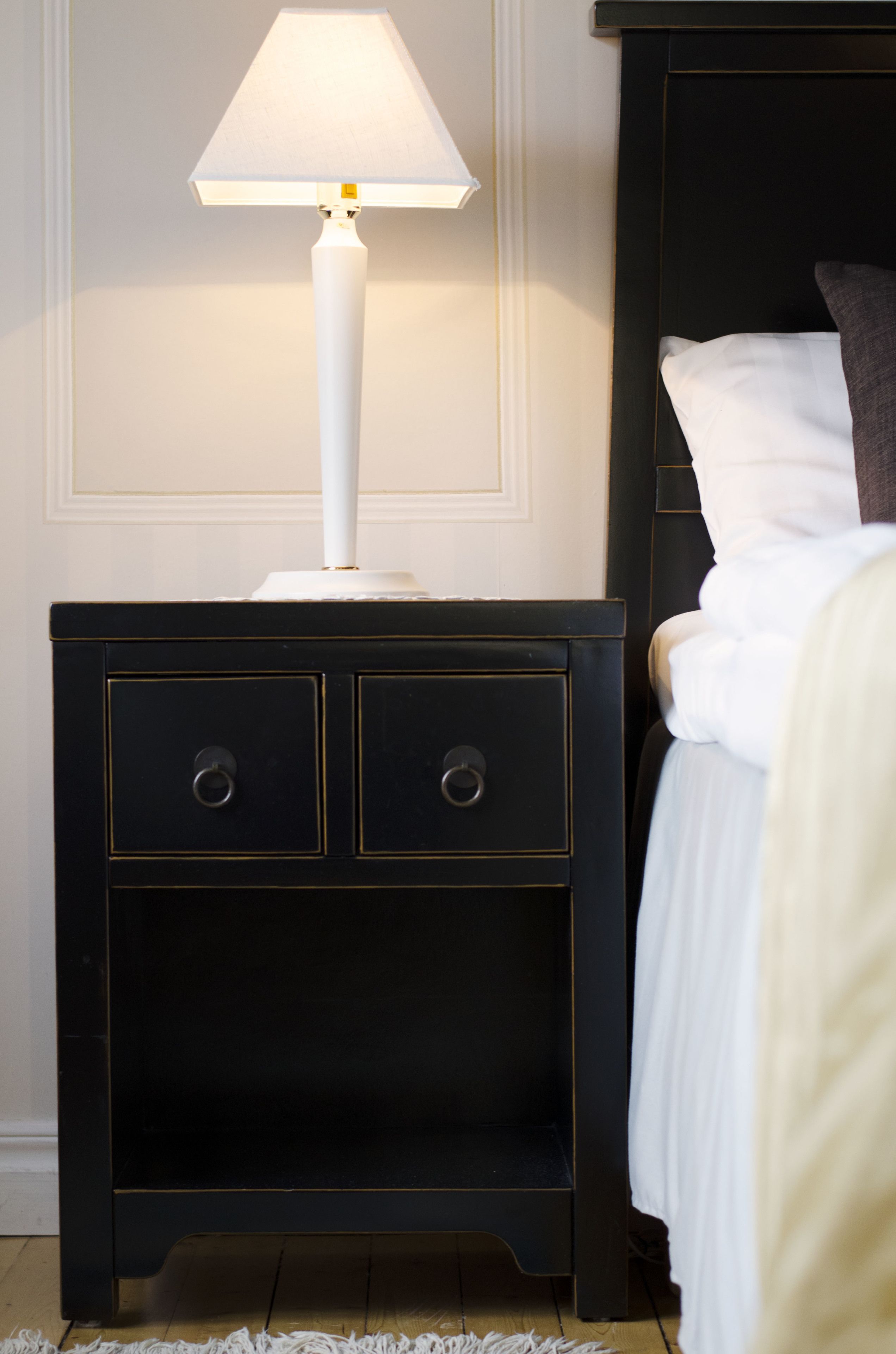 Un comodino con una lampada bianca, due cassetti e una testiera in legno scuro con un cuscino bianco.
