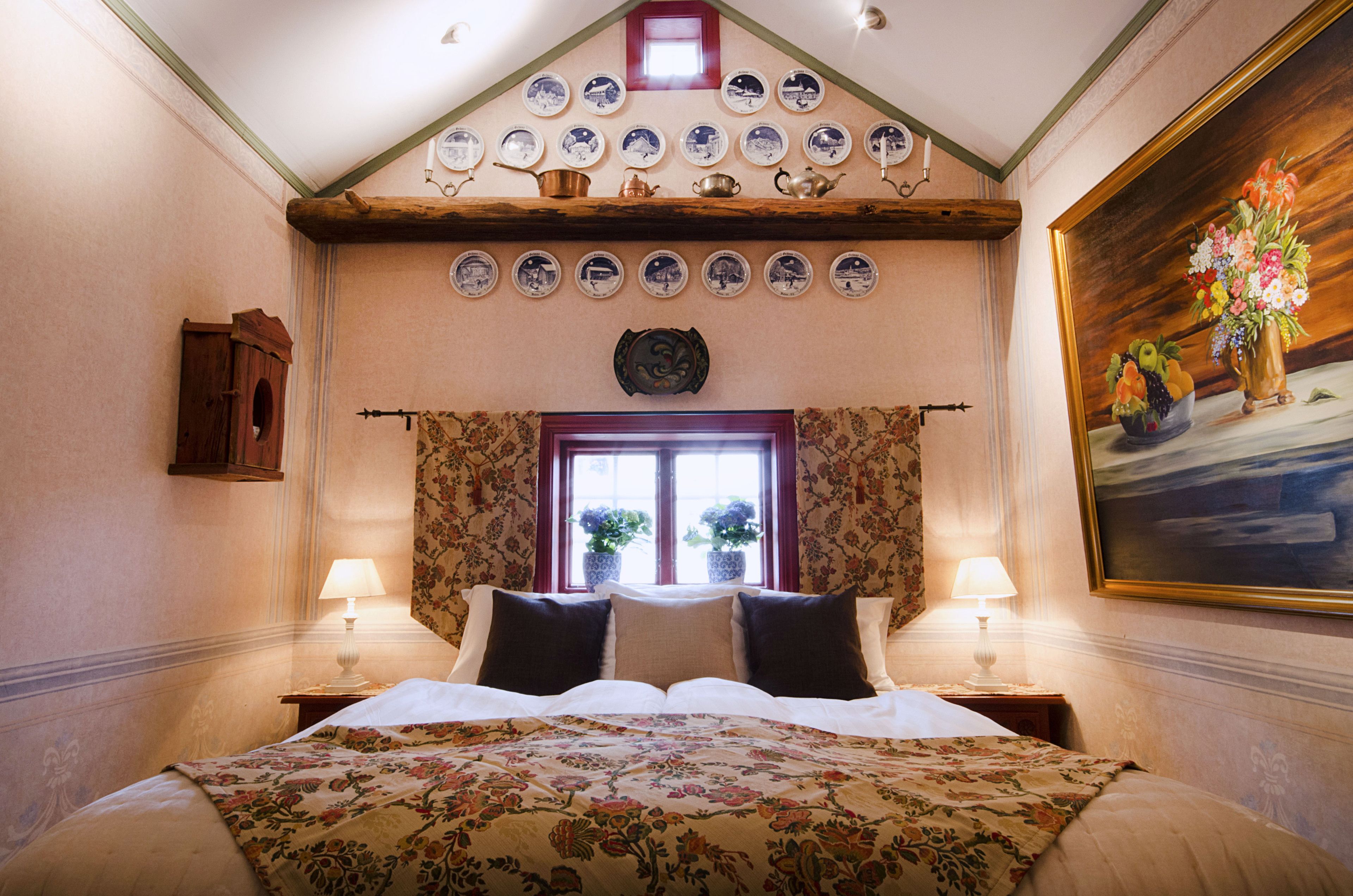 Un dormitorio con una cama grande, dos mesitas de noche con lámparas, una ventana con cortinas, un estante de pared con platos y un cuadro.