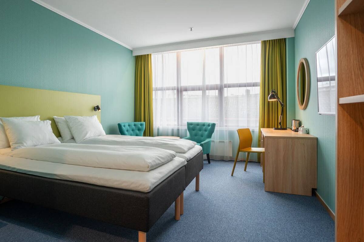 Une chambre d’hôtel avec un lit double, un bureau, deux chaises et une grande fenêtre avec des rideaux.