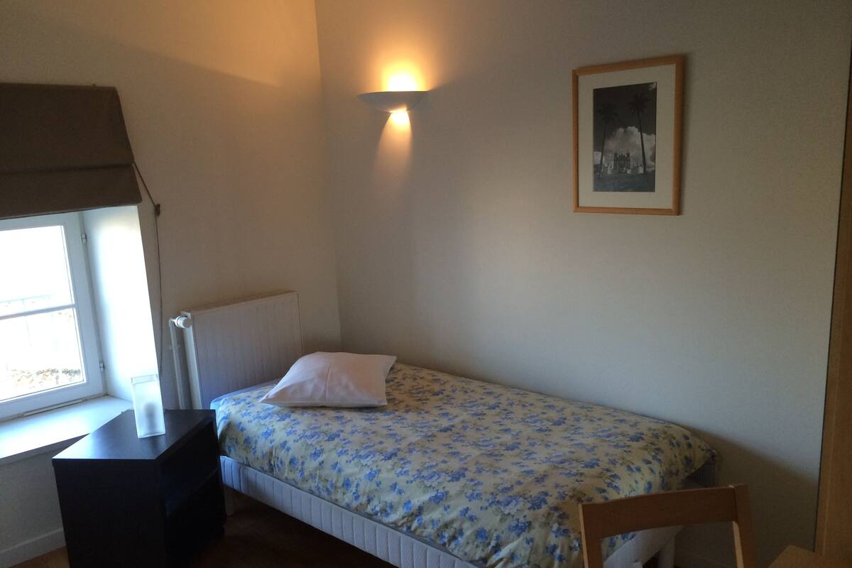 Ein kleines Zimmer mit einem Einzelbett, einem Fenster, einem Nachttisch, einem Heizkörper und einem gerahmten Bild an der Wand.