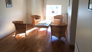 Una habitación con suelo de madera, sillas de mimbre, una mesa con superficie de vidrio y un cuadro en la pared.