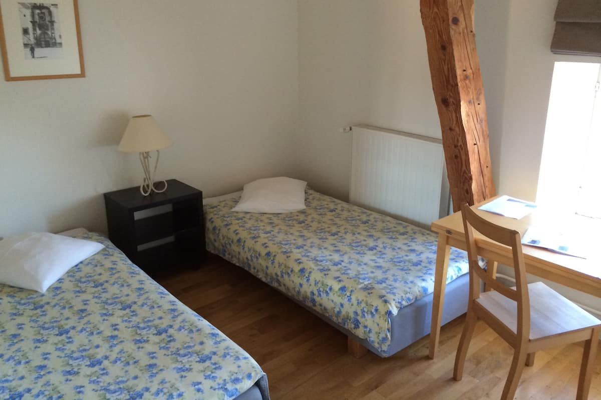 Ein kleiner, schlichter Raum mit zwei Einzelbetten, einem Holztisch, einem Stuhl, einem Nachttisch und einer Lampe.