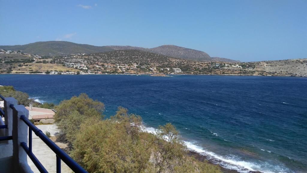 A coastal view with a clear blue sea, a hillside with buildings, and a railing in the foreground.