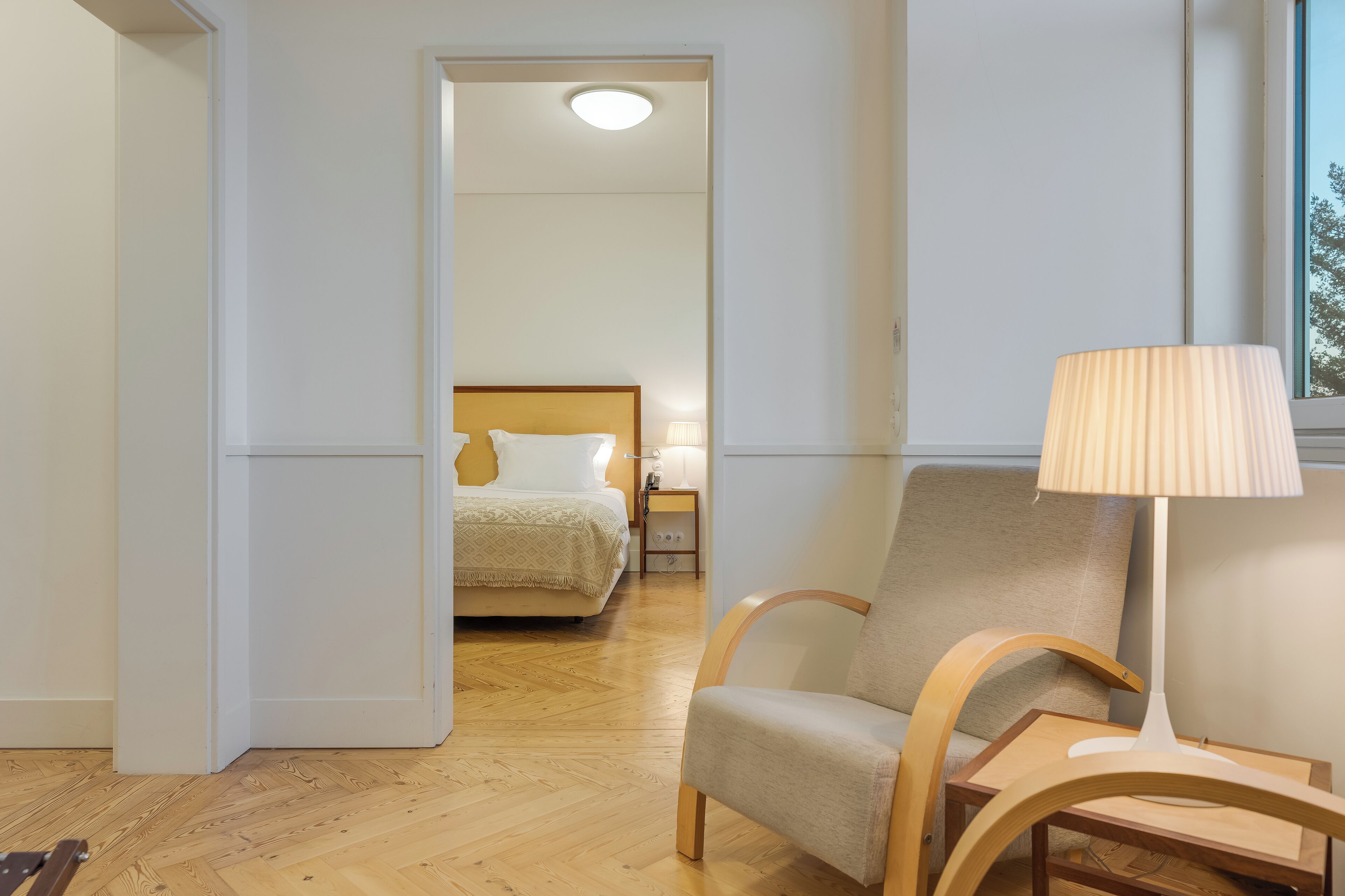 Una habitación de hotel con piso de madera, una cama con cabecera, una silla, una mesa y una lámpara.
