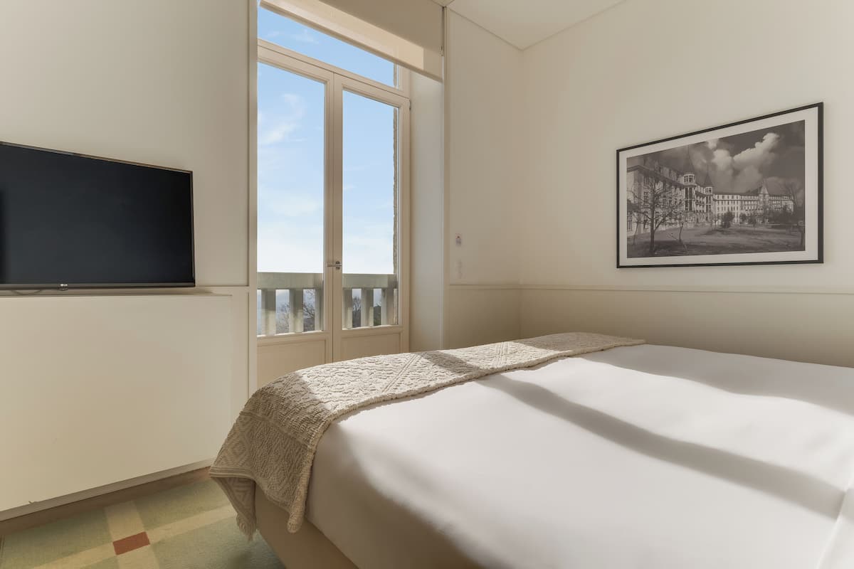 Een hotelkamer met een bed, een televisie aan de muur, een raam en een lijst met een foto.