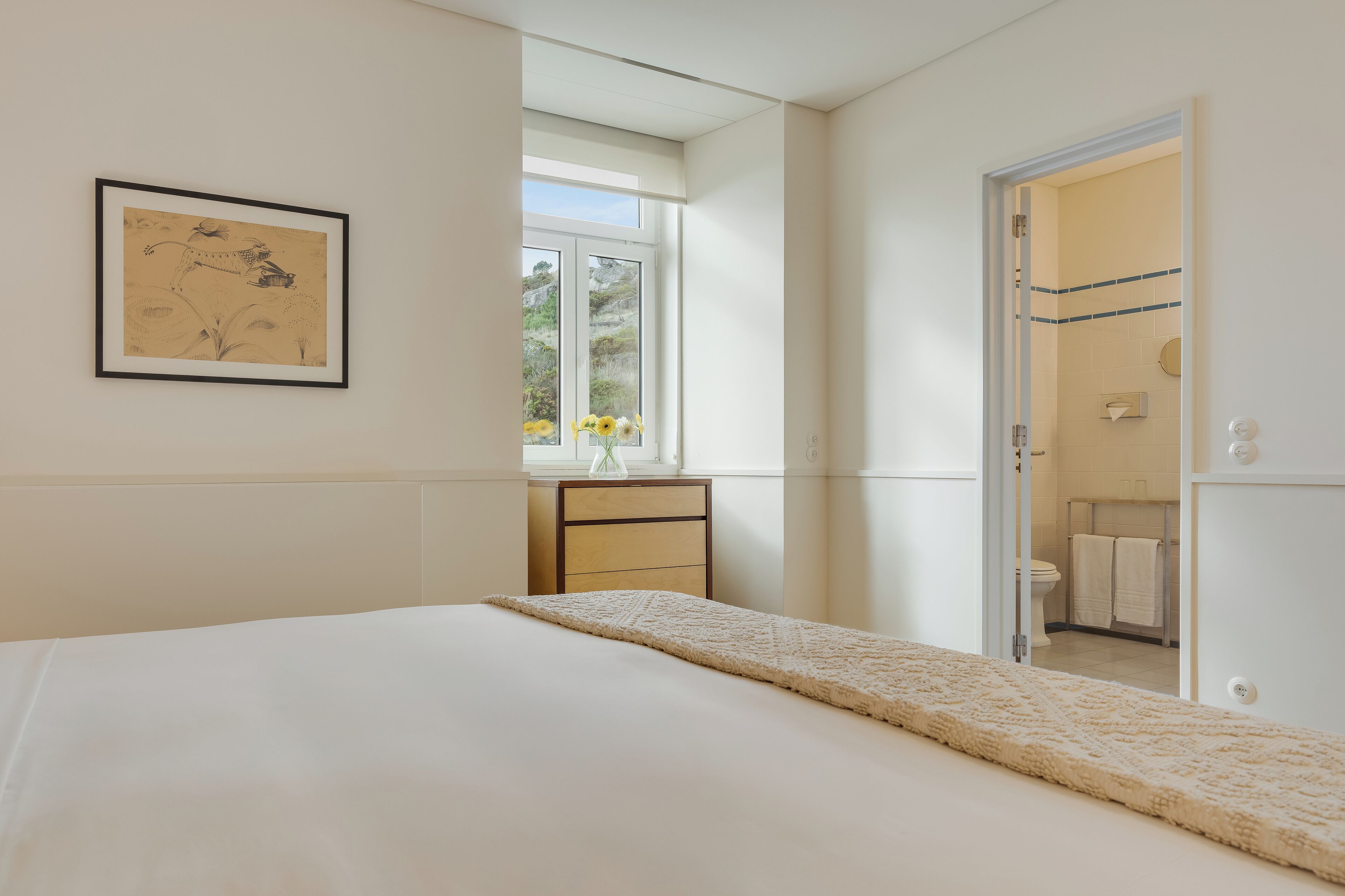 Une chambre à coucher avec un lit, une table de chevet, un tableau encadré au mur et une vue sur la salle de bain par une porte ouverte.