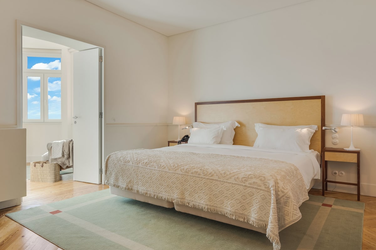 Een slaapkamer met een groot bed, bedlampjes, een nachtkastje en uitzicht op de lucht door een open deur.