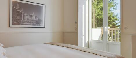 A hotel room with a large bed, a framed picture on the wall, and a view of a balcony with trees.