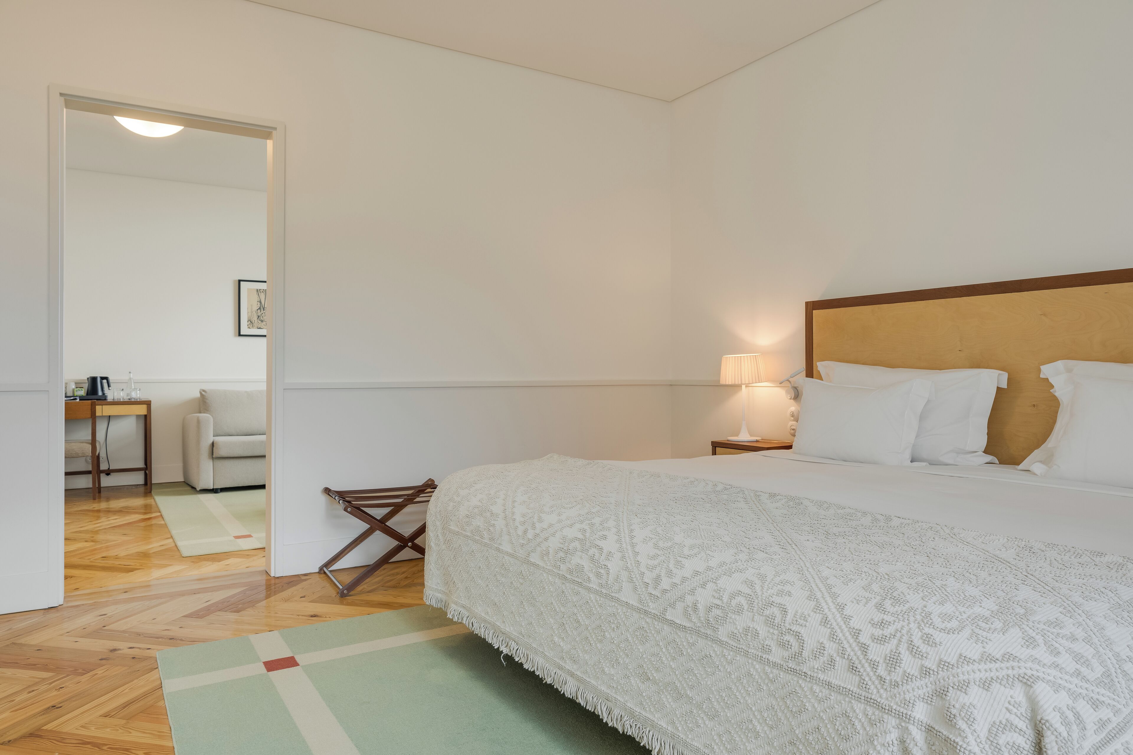 A bedroom with a large bed, a nightstand with a lamp, a wooden floor, and a view of a dining area through an open door.
