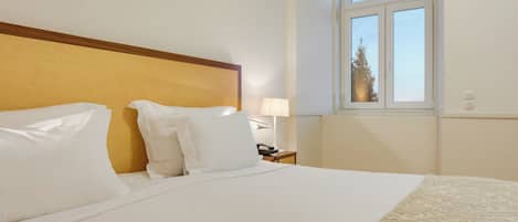 A hotel room with a bed, a nightstand with a lamp, and a window with a view of a tree.