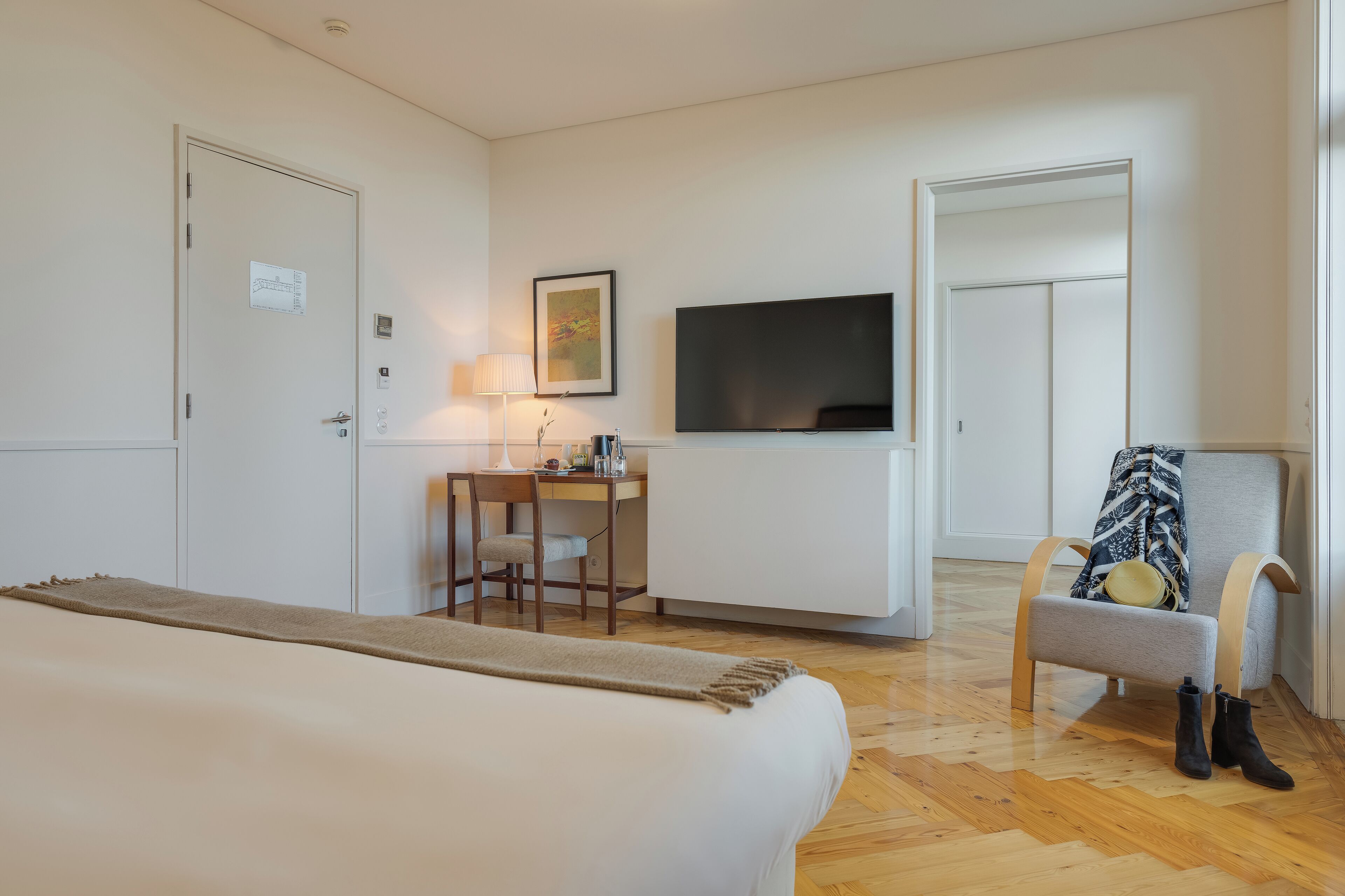 Una habitación de hotel con una cama, un escritorio con una silla, un televisor y un perchero con botas.