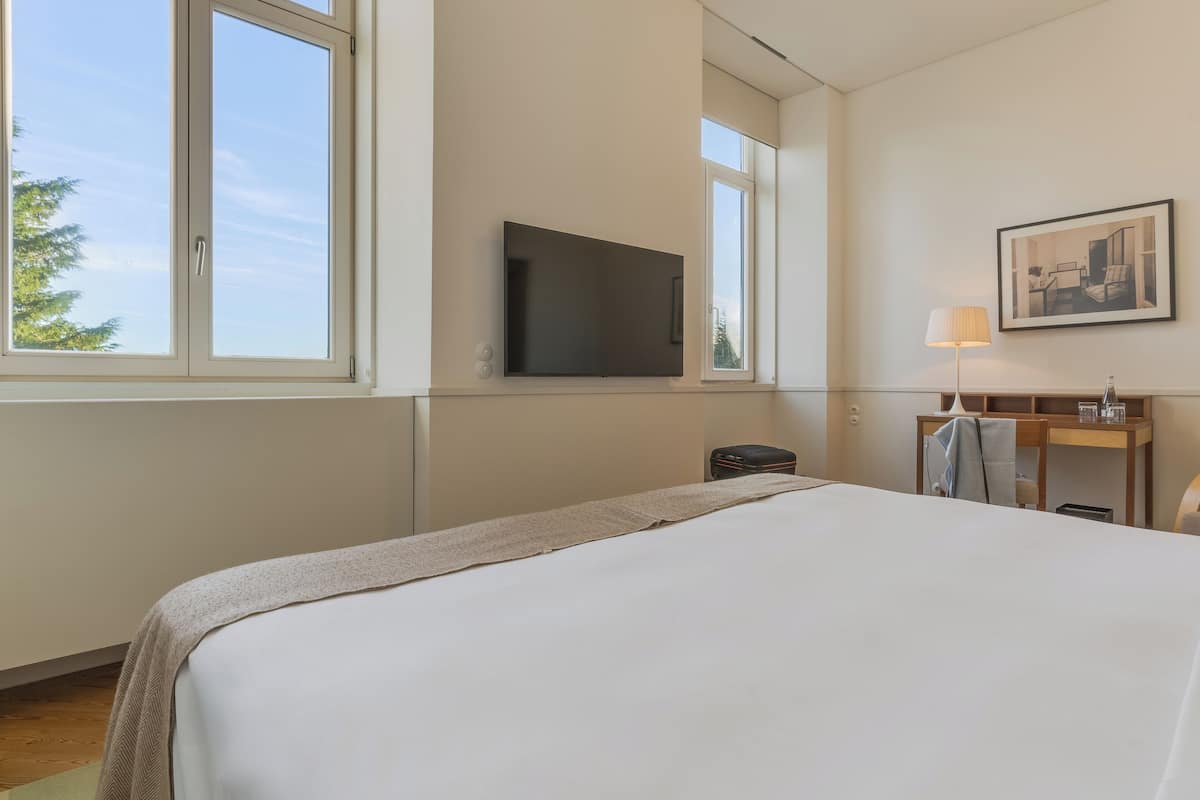 Een hotelkamer met een groot bed, een flatscreen televisie aan de wand, een bureau met een lamp en een raam met uitzicht op bomen.