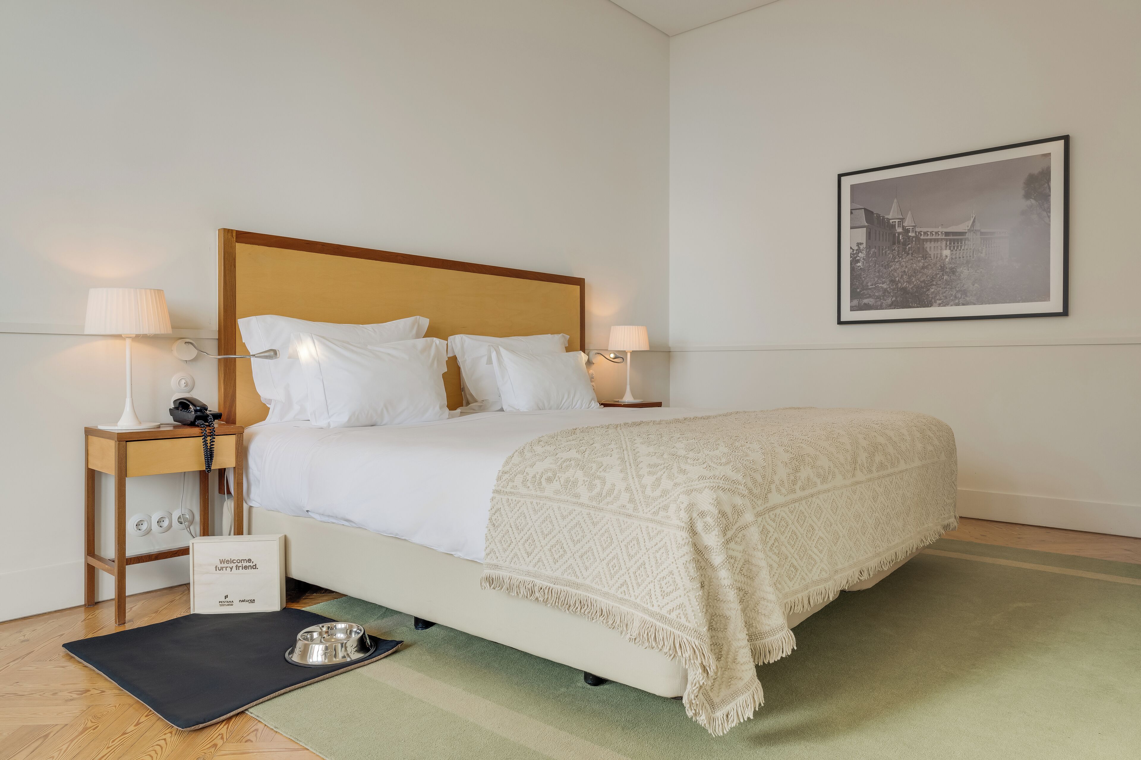 A neatly made bed with a wooden headboard, two bedside lamps, a framed picture on the wall, and a welcome sign on the floor.