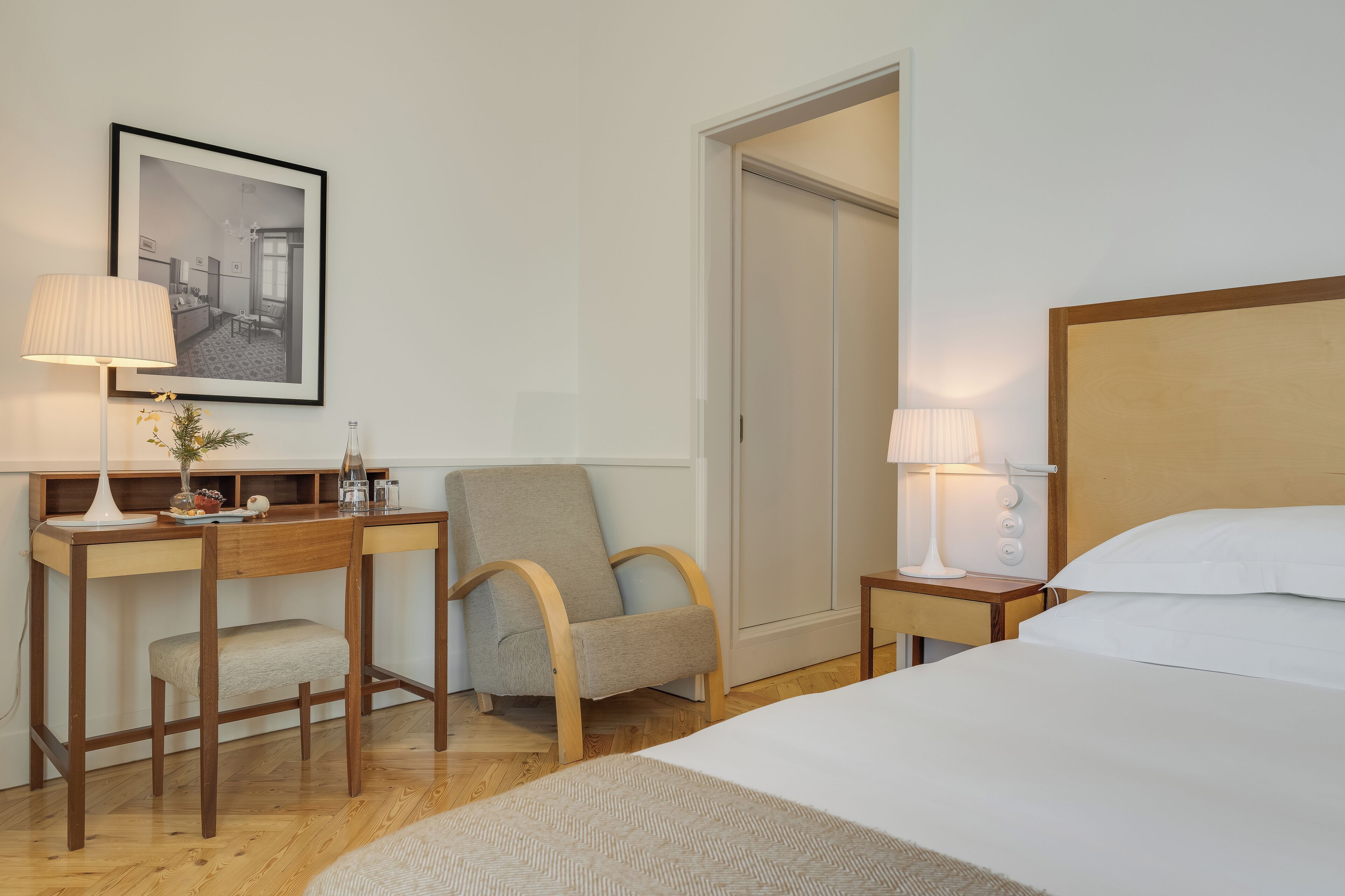 A hotel room with a bed, a desk with a lamp, a chair, and a framed picture on the wall.