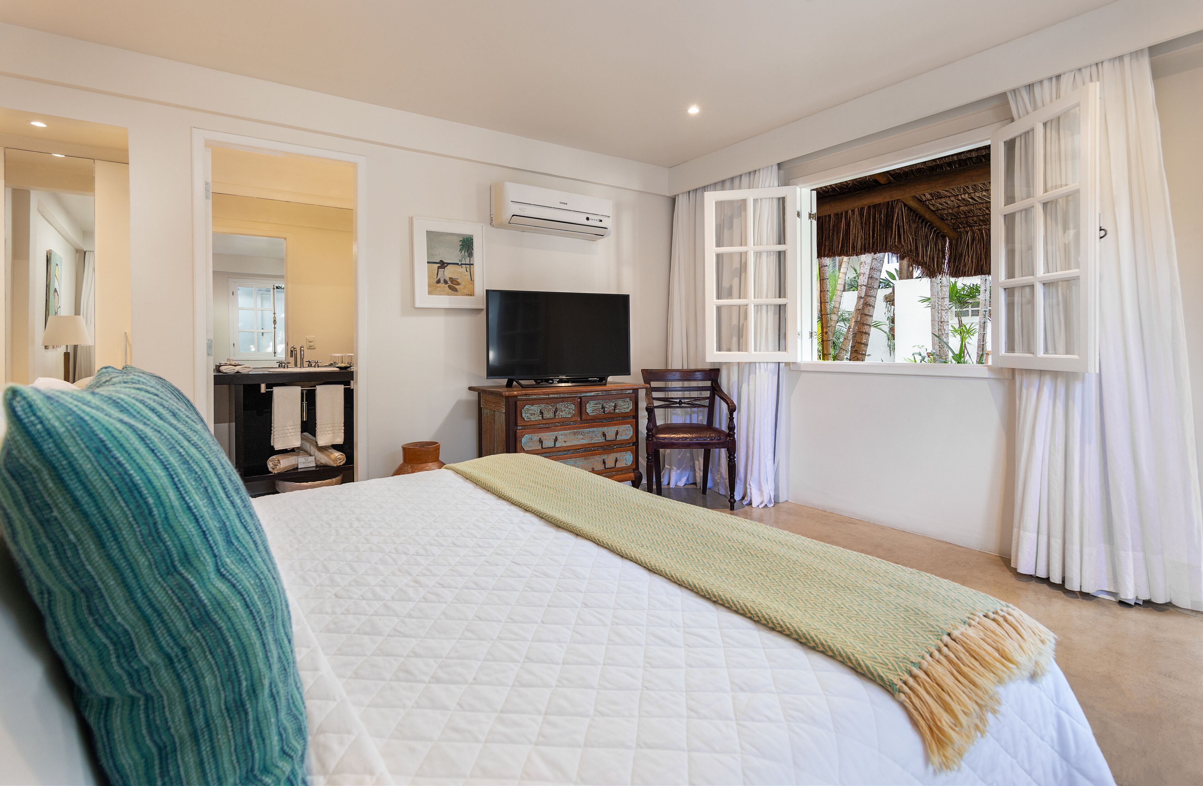 A bedroom with a large bed, a TV, a wooden dresser, a chair, and a window with curtains.