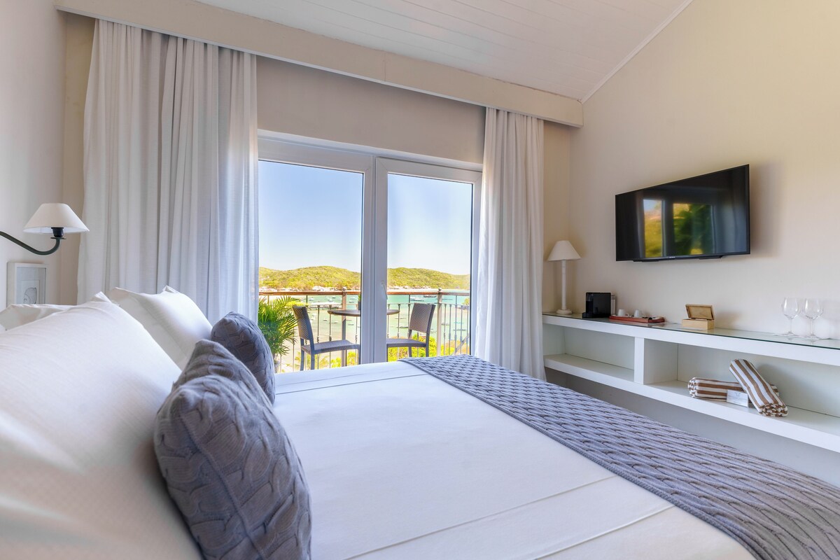 A hotel room with a large bed, a TV mounted on the wall, a balcony with a view of the hills, and a small desk with a glass top.