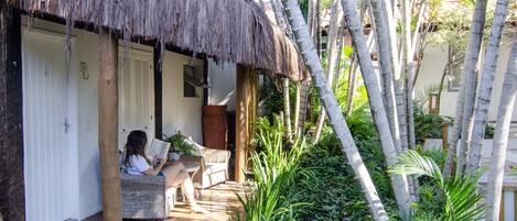 A thatched-roof veranda with a person reading a book, surrounded by lush greenery and palm trees.