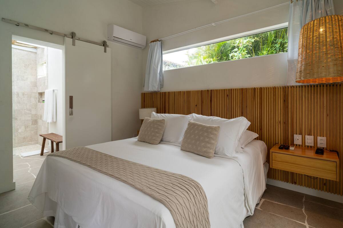 Un dormitorio con una cama grande, cabecera de madera, mesita de noche con lámpara y ventana con vista a zonas verdes.